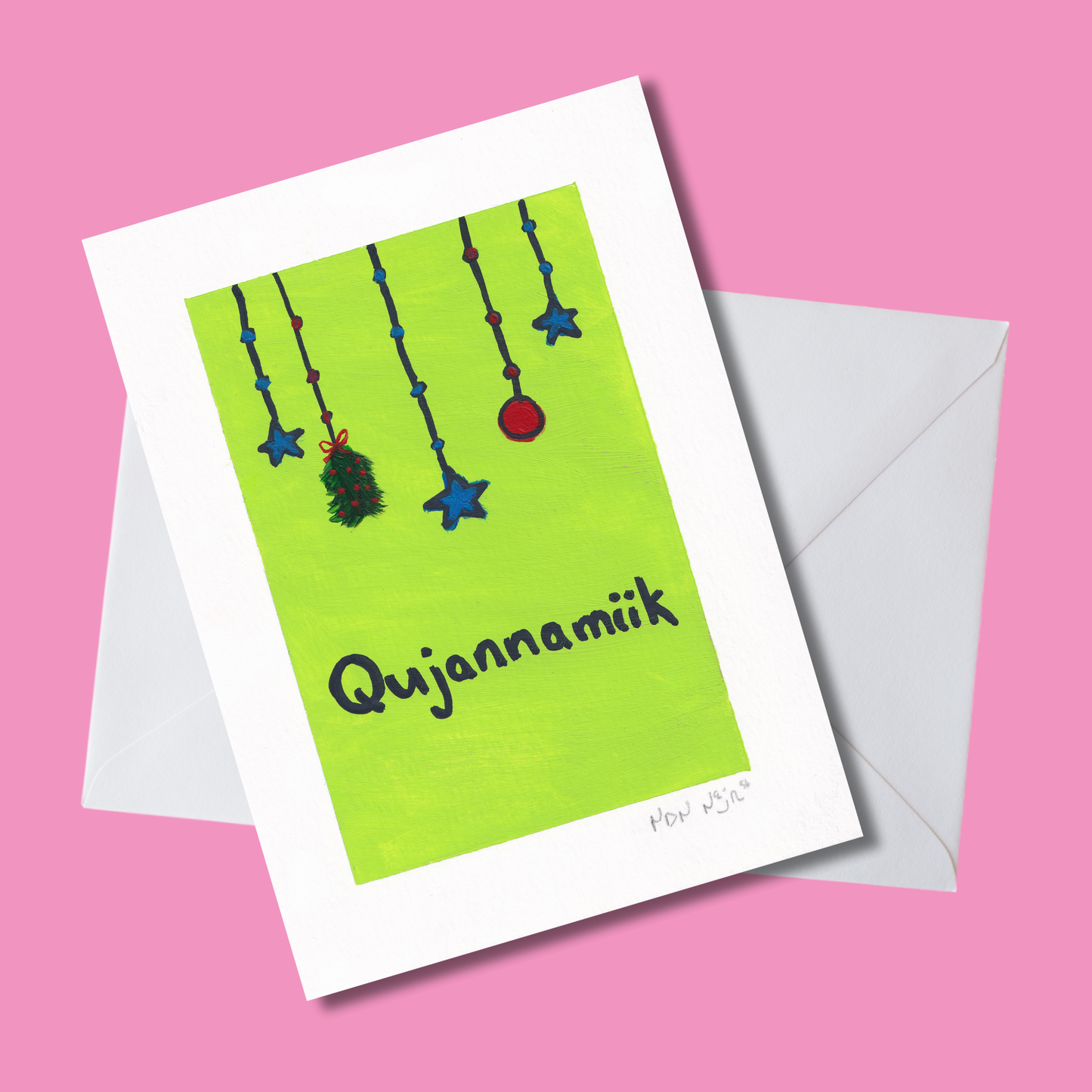 Hand-painted ‘Qujannamiik’ greeting card on a bright pink background with white envelope, featuring hanging blue stars, a red ornament, and a green sprig above the word meaning ‘thank you’ in Inuktitut.