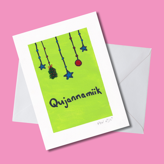 Hand-painted ‘Qujannamiik’ greeting card on a bright pink background with white envelope, featuring hanging blue stars, a red ornament, and a green sprig above the word meaning ‘thank you’ in Inuktitut.
