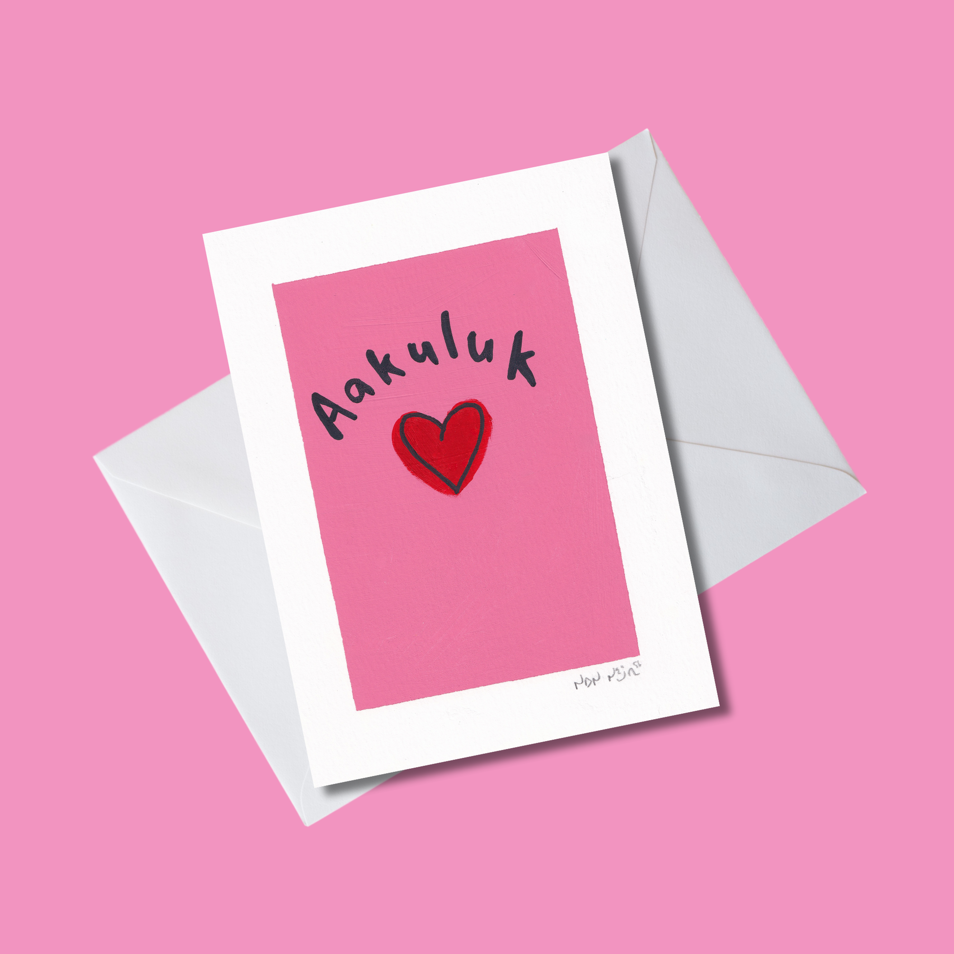 Angled view of a hand-painted artwork titled ‘Aakuluk,’ featuring a bold red heart centered on a bright pink background/white envelope with the Inuktitut word ‘Aakuluk,’ meaning ‘I love you,’ written above in black. The artwork rests on a soft pink surface, radiating warmth, simplicity, and affection.